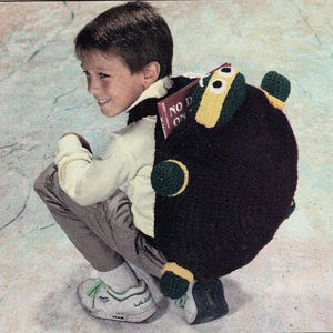 May include: A dark brown crocheted backpack with green and yellow detailing. The backpack has a unique, round shape and a playful design. A book with the text "NO D ON" is visible. The backpack is being worn by a person.