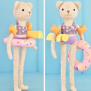 May include: A beige bear doll wearing a purple floral swimsuit, orange arm floaties, and a pink inflatable ring. The doll is holding a yellow rolled-up towel. The background is a light blue.