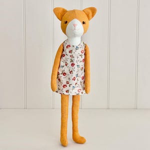Könnte beinhalten: Eine handgemachte orangefarbene Katzendolle, die ein weißes Kleid mit Blumenmuster trägt. Die Puppe hat ein weißes Gesicht mit schwarzen Augen und einer rosa Nase.