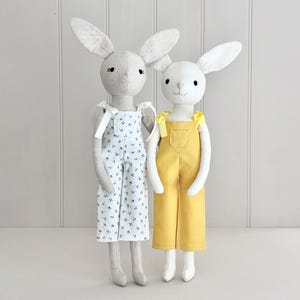 以下が含まれることがあります： 手作りのぬいぐるみウサギのドール2体。1体は花柄の白いオーバーオール、もう1体は黄色のオーバーオールを着ています。