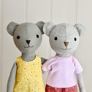 Peut inclure: Deux ours en tissu gris portant des tenues différentes. Un ours porte une robe jaune à motif floral. L'autre ours porte une chemise rose et un pantalon à motif rouge et blanc.