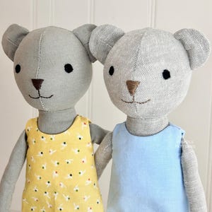以下が含まれることがあります： 異なる服装をした2体のグレーのリネン製のテディベア。1体のベアは白のドット柄の黄色のドレスを着ています。もう1体のベアは薄いブルーの袖なしのドレスを着ています。