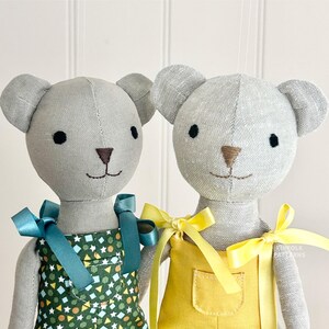 Op de afbeelding: Twee grijze teddyberen van linnen met overalls. Eén beer draagt een tealblauwe overall met een geometrisch patroon in groen, wit en geel. De andere beer draagt een gele overall. Beide beren hebben een bruine geborduurde neus en zwarte ogen.