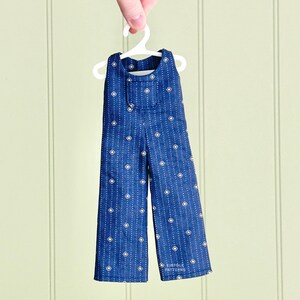 以下が含まれることがあります： 白いハンガーに掛けられた、白いダイヤモンドとドット模様の青いジャンプスーツ。ジャンプスーツには胸に小さなポケットとワイドレッグが付いています。「KINFOLK PATTERNS」の文字が確認できます。