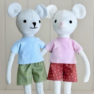 以下が含まれることがあります： 服を着たぬいぐるみネズミ2体。左側のネズミは薄い青色のTシャツと緑色のショートパンツを着用しています。右側のネズミはピンク色のTシャツと白い花柄の赤いショートパンツを着用しています。