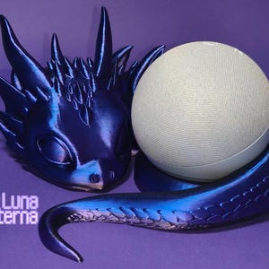 Könnte beinhalten: Ein 3D-gedruckter Drache, der sich um einen weißen intelligenten Lautsprecher schlingt. Der Drache ist tiefblau mit einem Farbverlauf zu hellblau an seinem Schwanz. Die Augen des Drachen sind geschlossen und er scheint zu schlafen. Der Lautsprecher ist ein rundes, weißes Gerät mit einer Stoffabdeckung.