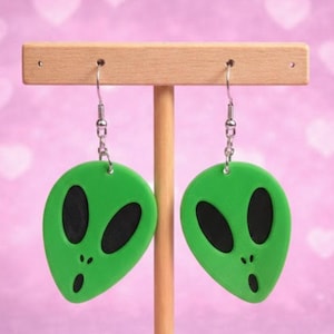 May include: A pair of green alien-shaped earrings with black oval eyes and a small open mouth. The earrings are suspended from silver-colored fish hook ear wires. The earrings are displayed on a wooden earring holder against a pink background.
