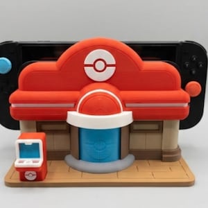Può includere: Un supporto a tema Pokémon Center, stampato in 3D, rosso e bianco per una Nintendo Switch. Il supporto presenta un design dettagliato con un tetto rosso, una porta blu e una piccola macchina arcade. La Nintendo Switch è nera.