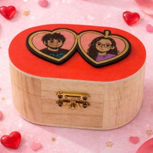 Può includere: Un portagioie in legno con coperchio rosso e chiusura dorata. La parte superiore presenta due ritratti a forma di cuore di una coppia. La scatola è realizzata in legno chiaro e decorata con piccoli cuori.