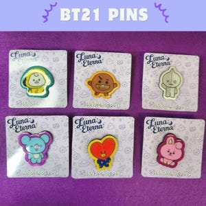 May include: Six colorful BT21 pins on cards. Each pin features a different cartoon character design with bright colors. The cards are white with the text "Luna Eterna" and "HANDMADE PINS". The background is a solid purple.