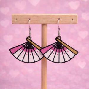 May include: A pair of dangle earrings featuring a fan design. The earrings have a pink and white fan with black outlines and a gold handle. The earrings are displayed on a wooden stand against a pink background.
