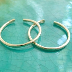 May include: Two gold-colored, open-ended hoop earrings. The earrings are circular with a small gap at the top. The earrings are displayed on a turquoise surface.