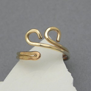 May include: A gold-toned metal ring featuring an open, adjustable band design. The top of the ring displays two symmetrical, curved loops that resemble a stylized heart shape. The metal surface has a hammered, textured finish throughout.