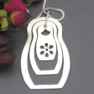 Pode incluir: Um colar com pingente de prata com um design de bonecas russas aninhadas. O pingente tem três camadas, com um recorte em forma de flor na boneca central. A corrente é fina, ideal para uso diário.