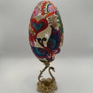 May include: A decorative egg with a colorful butterfly and heart design, made with rhinestones, on a gold swan stand.