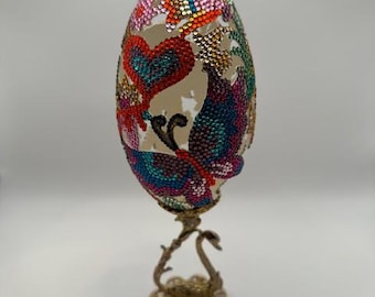 May include: A decorative egg with a colorful butterfly and heart design, made with rhinestones, on a gold swan stand.