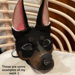 May include: A black and brown Doberman Pinscher mask with mesh eye coverings and a silver nose ring. The mask has pink inner ears and is displayed on a white surface. Text on the image reads "These are some examples of my work :)" and "Please note that I will not remake or duplicate any of them. They are not for sale!"