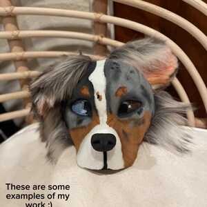 May include: A detailed dog mask with blue, brown, and gray fur, featuring a white stripe down the center of the face. The mask has realistic eyes and a black nose. Text on the image reads: "These are some examples of my work :)"