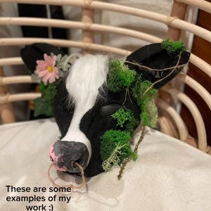 May include: A black and white cow head sculpture with a pink nose ring, adorned with a pink flower and green moss. The text on the image reads: "These are some examples of my work :) Please note that I will not remake or duplicate any of them. They are not for sale!"