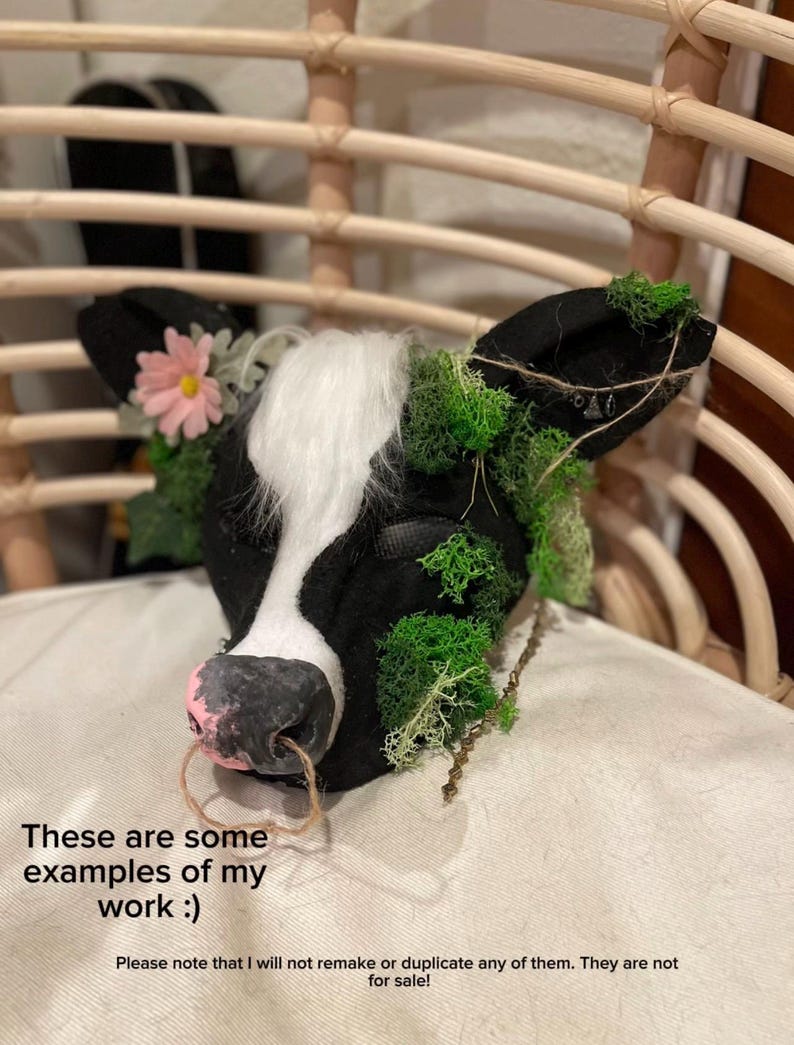 May include: A black and white cow head sculpture with a pink nose ring, adorned with a pink flower and green moss. The text on the image reads: "These are some examples of my work :) Please note that I will not remake or duplicate any of them. They are not for sale!"