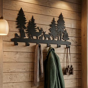 May include: Black metal wall-mounted coat rack featuring a forest scene with deer and trees. The rack has multiple hooks for hanging coats, scarves, and other items. A green coat and a scarf hang from the hooks. A bench below holds books and a bowl.
