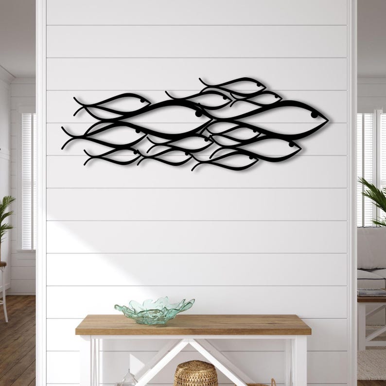 May include: A black metal wall art piece depicting a school of fish swimming together. The fish are stylized with curved lines and eye details. The artwork is mounted on a white shiplap wall, creating a coastal theme.