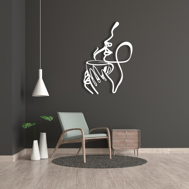 K&ouml;nnte beinhalten: Wei&szlig;e Wandkunst aus Metall, die ein stilisiertes Frauen Gesicht mit einer Hand, die eine Tasse Kaffee h&auml;lt, darstellt. Die Kunst ist an einer dunkelgrauen Wand in einem Wohnzimmer angebracht.