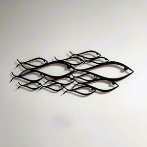 May include: Black metal wall art depicting a school of fish. The fish are stylized with curved lines and eye details, creating a flowing, aquatic design. The artwork is mounted on a white wall, offering a contrast that highlights the fish's forms.
