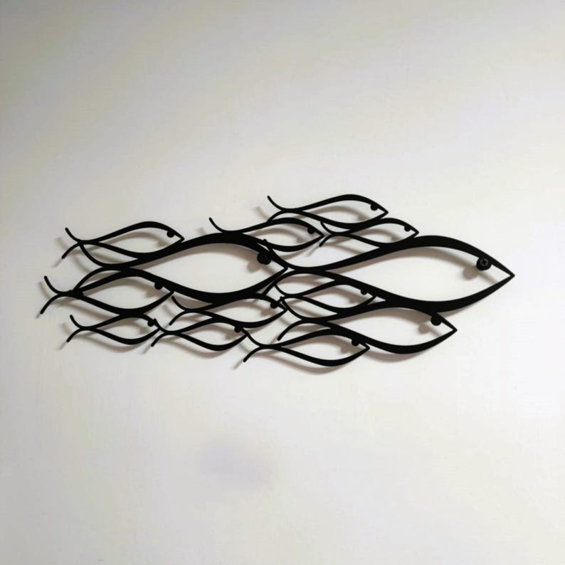 May include: Black metal wall art depicting a school of fish. The fish are stylized with curved lines and eye details, creating a flowing, aquatic design. The artwork is mounted on a white wall, offering a contrast that highlights the fish's forms.