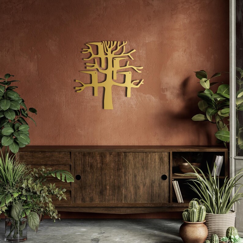 May include: A gold-colored metal tree wall art decor. The tree has a stylized design with branches and a trunk. The wall art is mounted on a textured, reddish-brown wall above a wooden cabinet.