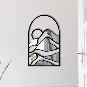 May include: Black metal arch-shaped wall mirror with a stylized mountain and sun design. The mirror reflects a view of a window and trees.