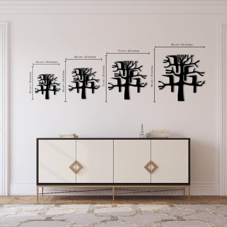 May include: Four black metal tree wall art pieces of varying sizes are displayed against a white wall. The sizes are 18 inches, 24 inches, 30 inches, and 35 inches. The art is above a white cabinet with gold accents.