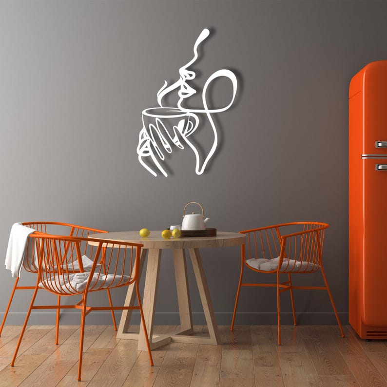 K&ouml;nnte beinhalten: Wei&szlig;e Wandkunst aus Metall, die eine stilisierte Frau darstellt, die eine Tasse Kaffee genie&szlig;t. Die Kunst ist an einer grauen Wand &uuml;ber einem Holztisch mit zwei orangefarbenen Metallst&uuml;hlen angebracht.