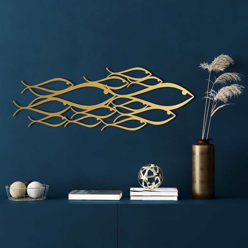 May include: A gold-colored metal wall art piece depicting a school of fish swimming together. The artwork is mounted on a dark blue wall. A vase with tall, dried plants and decorative objects are on a dark blue surface below the artwork.