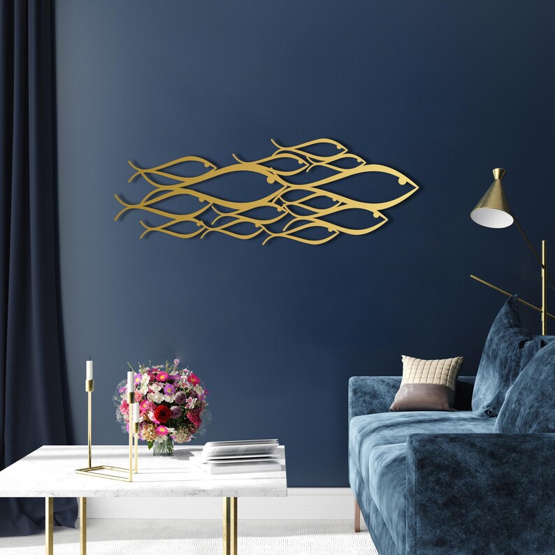 May include: A gold-colored metal wall art piece depicting a school of fish swimming together. The artwork is mounted on a dark blue wall, creating a striking contrast. The fish are stylized with flowing lines.