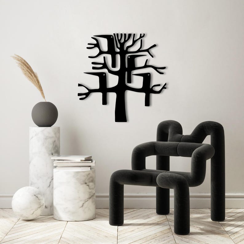 May include: A black metal wall art piece depicting a stylized tree with birds perched on its branches. The artwork is mounted on a white wall. A modern black chair and decorative items are in the foreground.