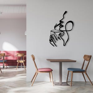 K&ouml;nnte beinhalten: Schwarze Wandkunst aus Metall, die eine stilisierte Frau darstellt, die eine Kaffeetasse mit aufsteigendem Dampf h&auml;lt. Die Kunst ist an einer wei&szlig;en Wand in einem Caf&eacute;-Ambiente mit Tisch und St&uuml;hlen angebracht.