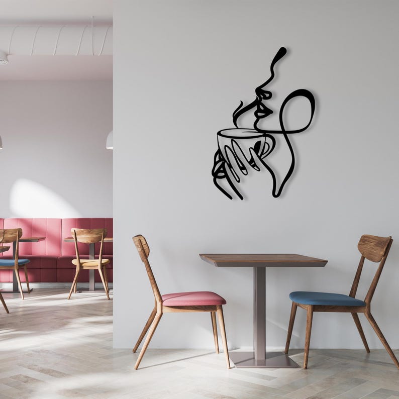 K&ouml;nnte beinhalten: Schwarze Wandkunst aus Metall, die eine stilisierte Frau darstellt, die eine Kaffeetasse mit aufsteigendem Dampf h&auml;lt. Die Kunst ist an einer wei&szlig;en Wand in einem Caf&eacute;-Ambiente mit Tisch und St&uuml;hlen angebracht.