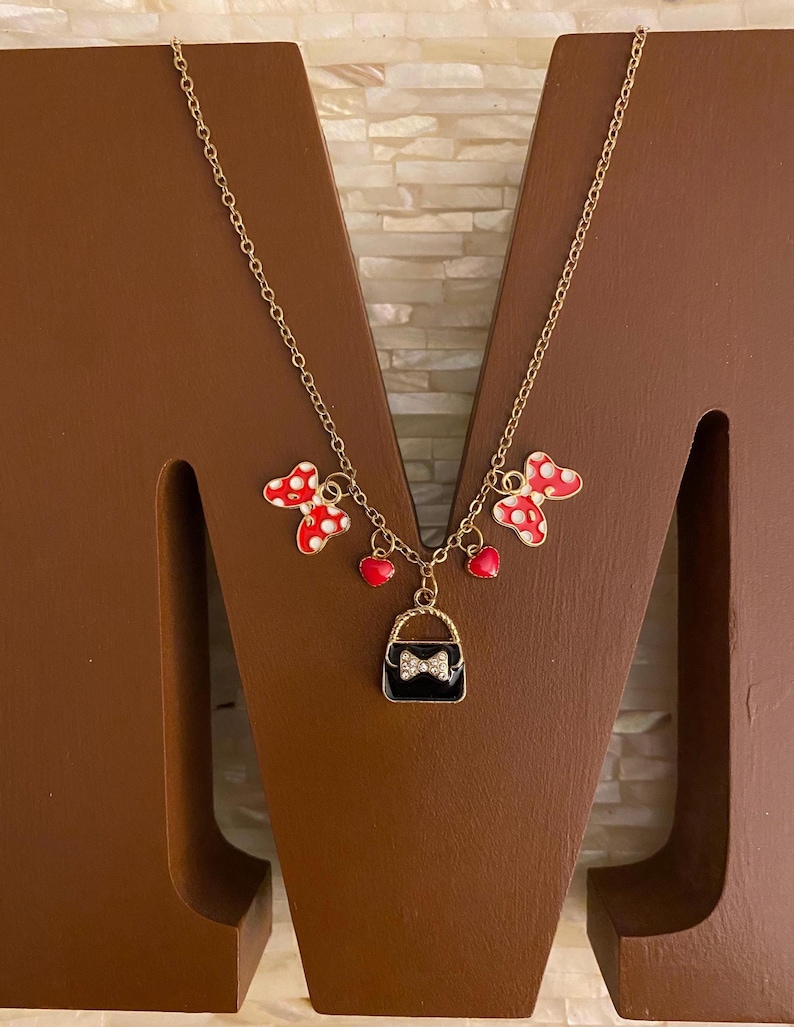 May include: Gold chain necklace with red and white polka dot bows, red hearts, and a black purse charm with a rhinestone bow.