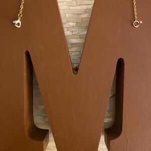 May include: A brown wooden letter M with two gold chains hanging from the top. The chains have a small, clear crystal at the end.