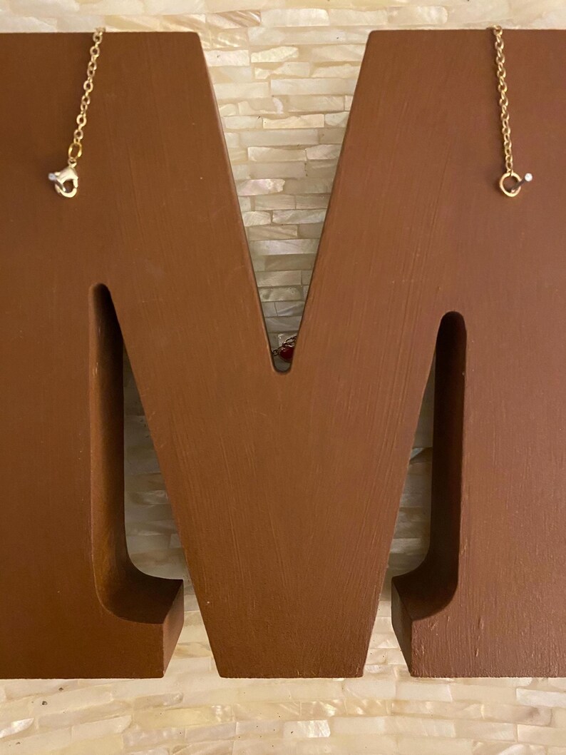 May include: A brown wooden letter M with two gold chains hanging from the top. The chains have a small, clear crystal at the end.