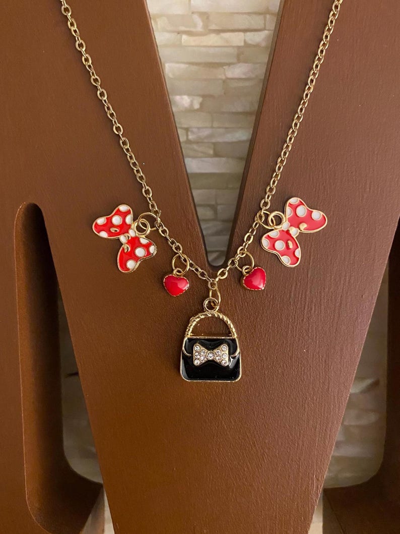 May include: Gold chain necklace with red and white polka dot bows, red heart charms, and a black purse charm with a rhinestone bow.