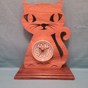 May include: A wooden cat clock with a gold-rimmed clock face. The clock face has black Roman numerals and black hands. The cat is brown and has a carved design.