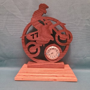 May include: A wooden clock featuring a silhouette of a motocross rider on a dirt bike. The clock face is round and silver with black numbers. The rider's number "77" is visible on the bike. The clock is mounted on a wooden base.