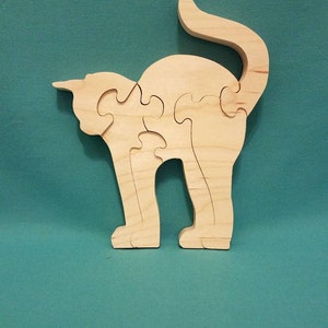 May include: A wooden cat puzzle, assembled and standing. The puzzle is made of light-colored wood and features a simple, stylized cat shape. The cat's tail is curved upwards, and the puzzle is on a teal background.