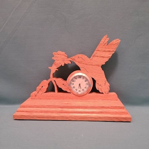 May include: A wooden clock featuring a hummingbird in flight, perched on a flower. The clock is on a wooden base and has a white face with black numbers.