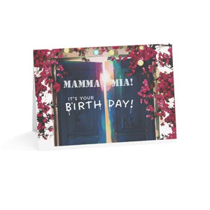 May include: A folded birthday card with the text "MAMMA MIA! IT'S YOUR BIRTHDAY!" in white. The card shows a blue door, bougainvillea flowers, and string lights. The design evokes a celebratory atmosphere.