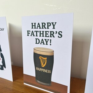 May include: A Father's Day greeting card with the text "HARPY FATHER'S DAY!" in dark green. The card features a pint of stout with a golden harp logo and the word "HAPPINESS". The card is on a wooden surface.