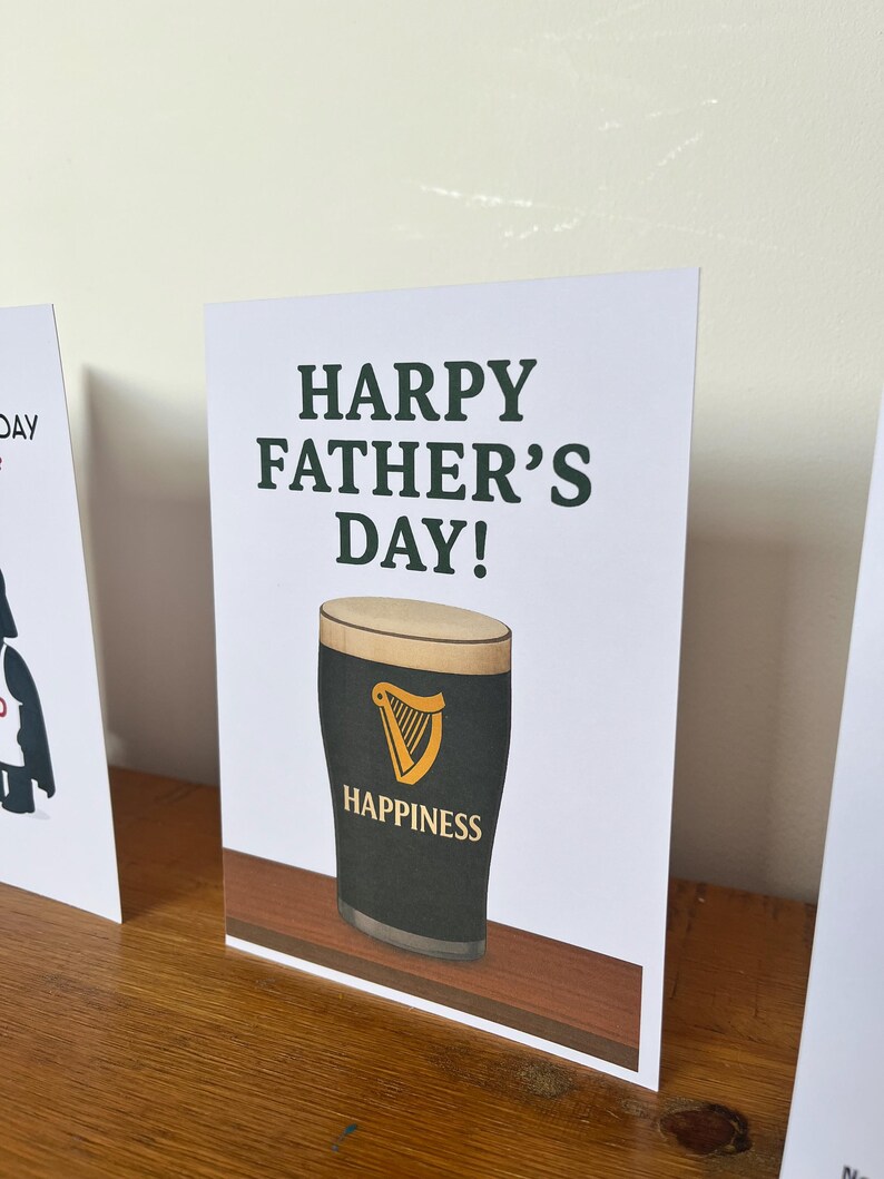 May include: A Father's Day greeting card with the text "HARPY FATHER'S DAY!" in dark green. The card features a pint of stout with a golden harp logo and the word "HAPPINESS". The card is on a wooden surface.