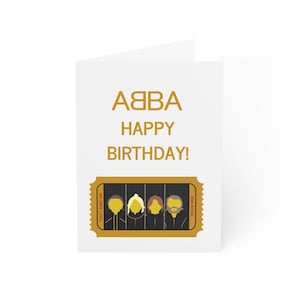 May include: A white card with gold text that reads "ABBA Happy Birthday!" and a gold ticket with a cartoon image of the band members.
